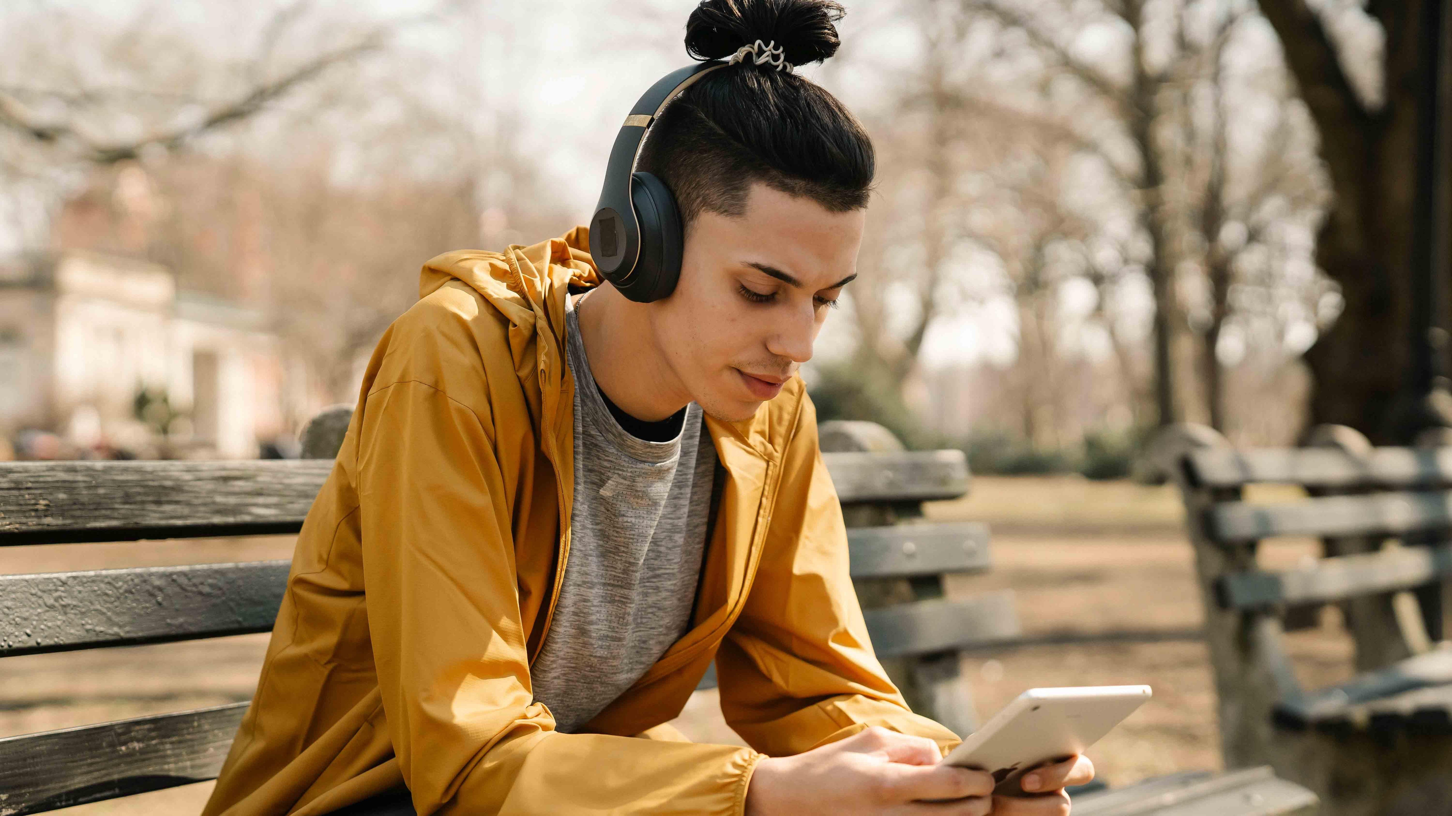Man wearing wireless headphones outdoors on park bench using tablet for immersive audio experience