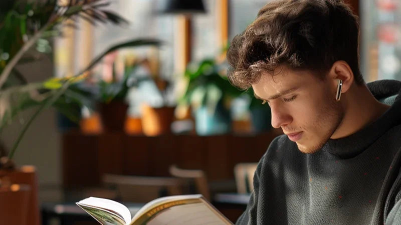 Man wearing wireless earbuds reading book in cozy cafe, enjoying immersive audio experience
