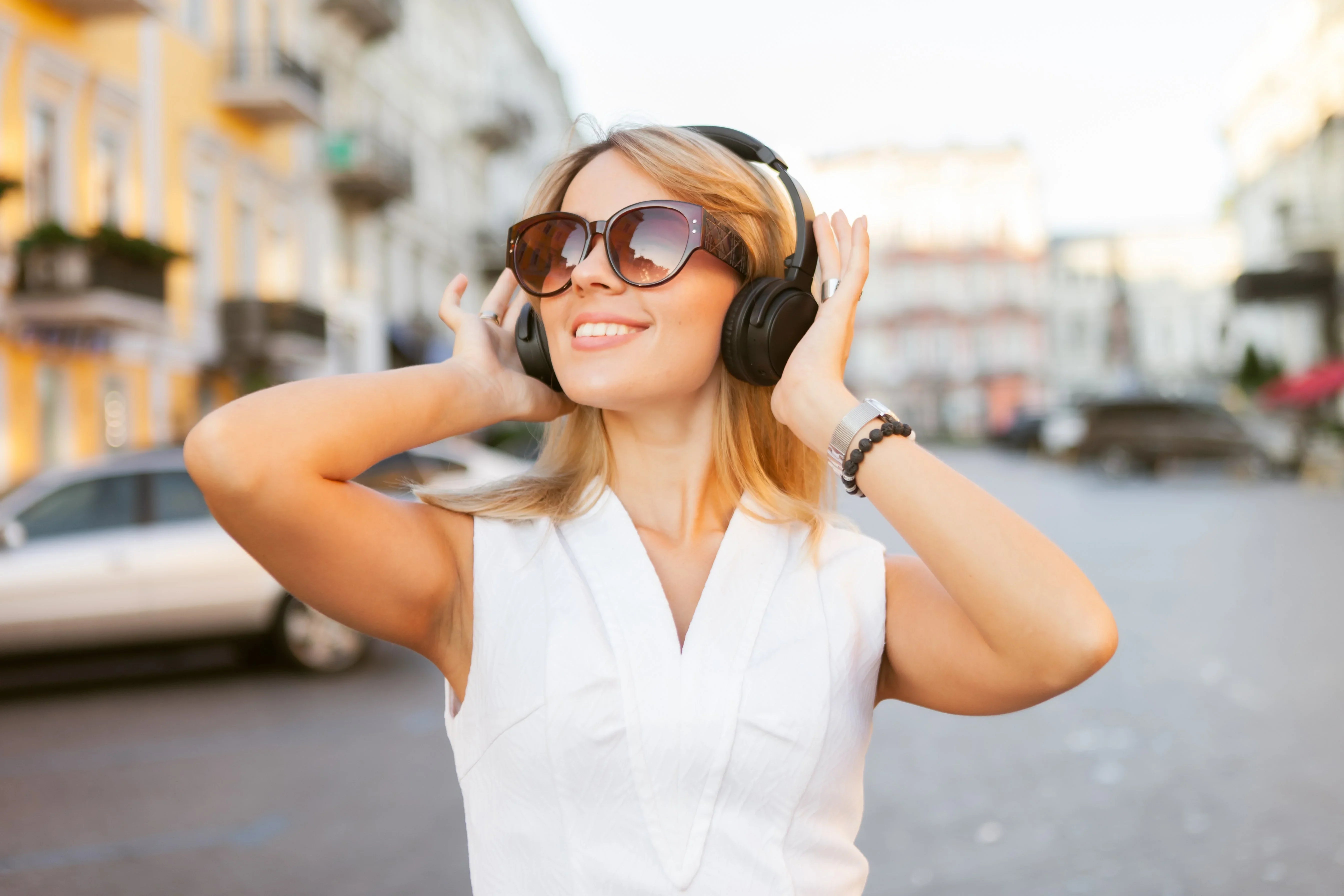 Woman enjoying music with wireless headphones on city street, stylish sunglasses, white top