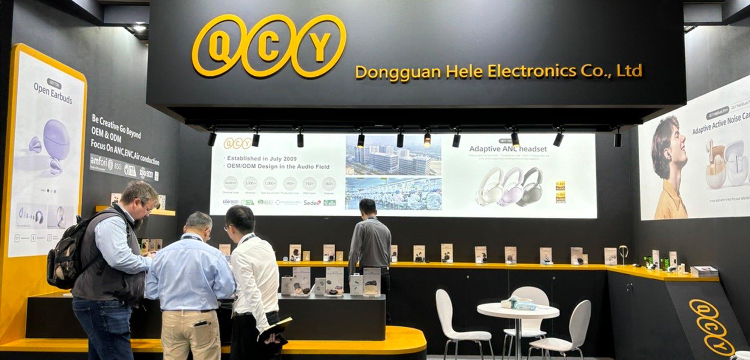 QCY booth at trade show displaying wireless earbuds and adaptive ANC headphones with visitors browsing products