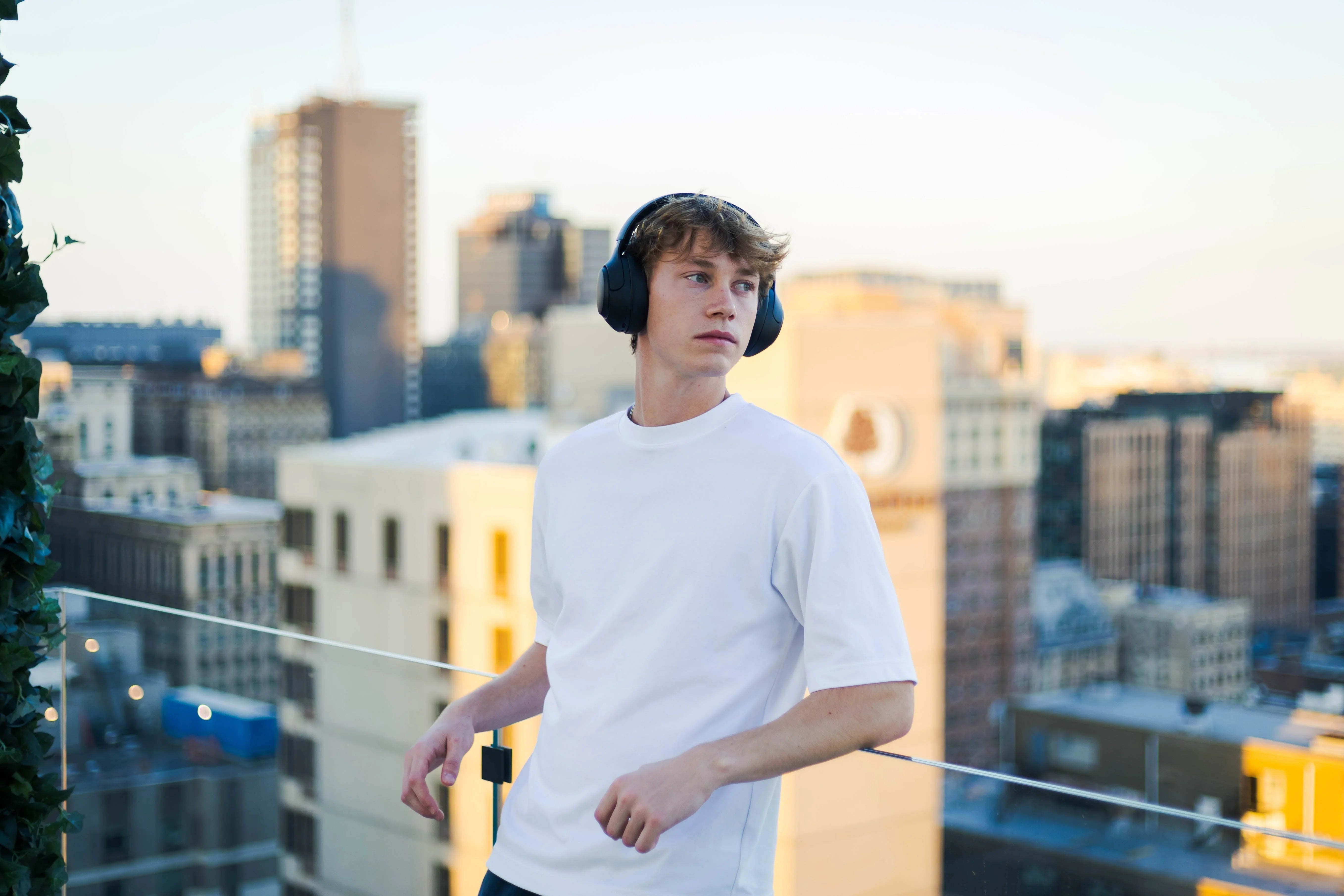 Young man wearing black wireless headphones on rooftop with city skyline, enjoying immersive audio experience