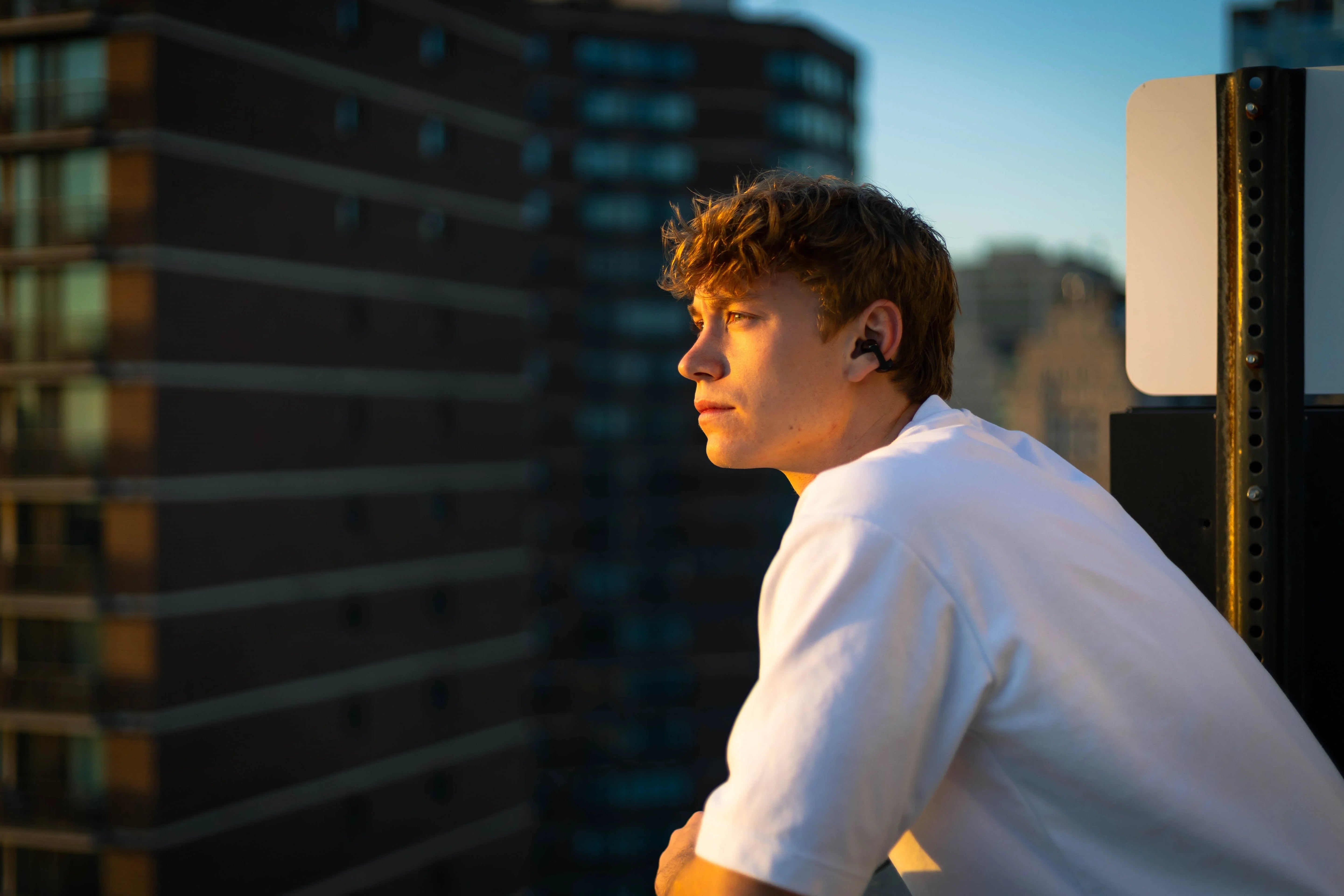 Young man wearing wireless earbuds on rooftop at sunset, city skyline background