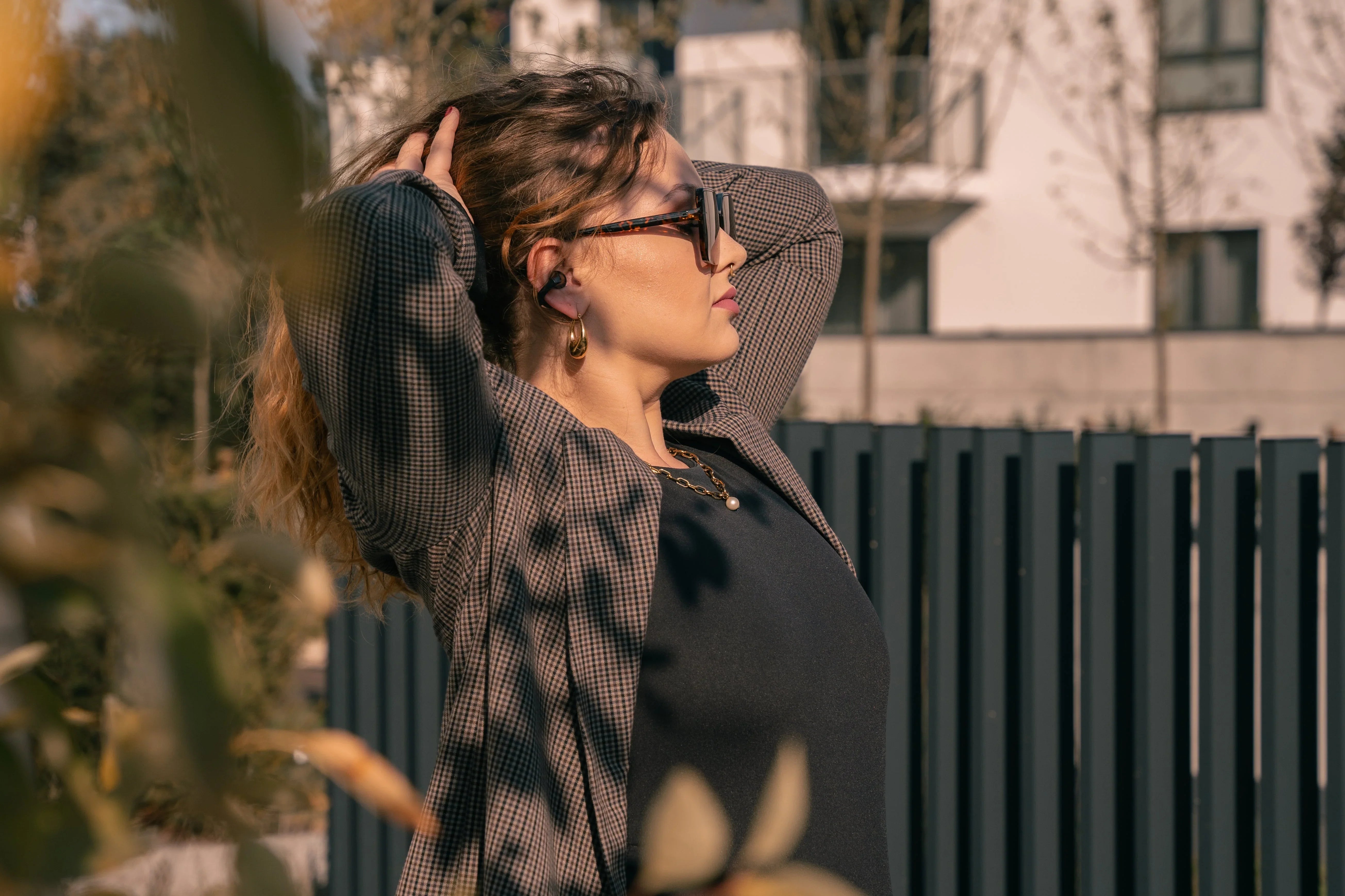Woman wearing wireless earbuds outdoors, enjoying music with sunglasses and stylish blazer