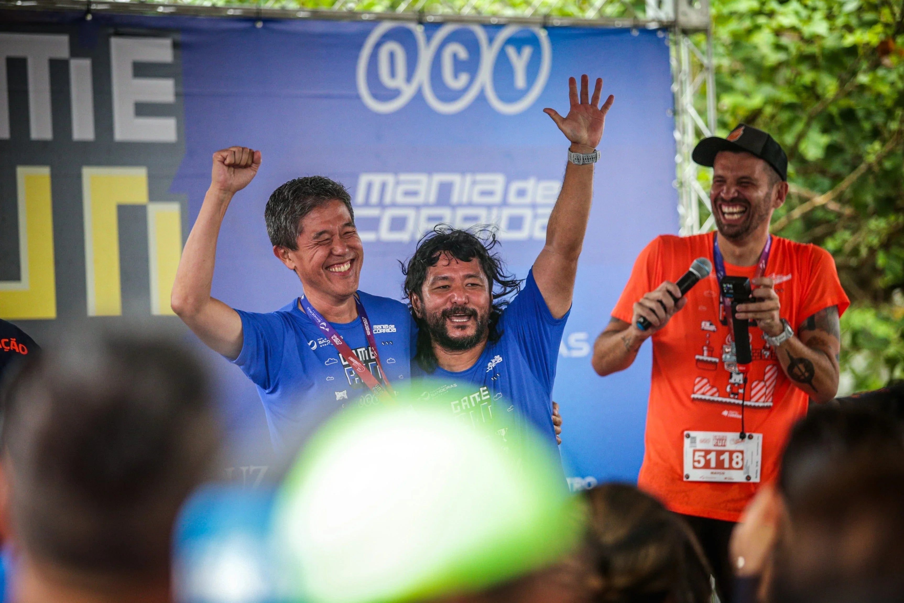 Men celebrating on stage at QCY Mania de Corrida event with medals and microphones
