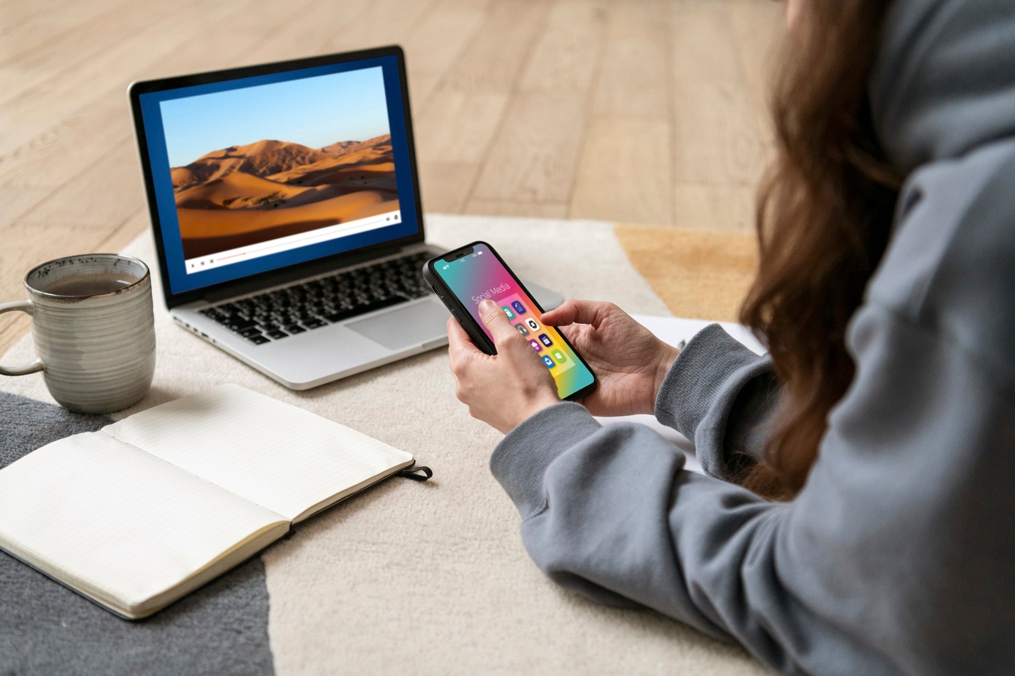 Person using smartphone and laptop on rug with coffee mug and notebook nearby