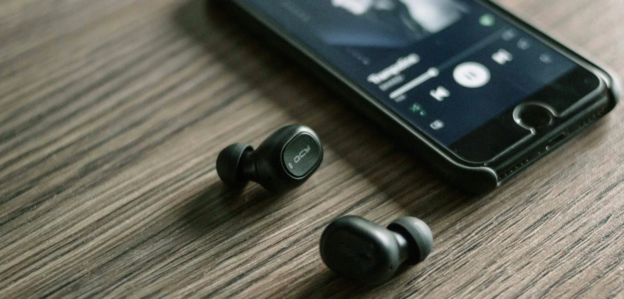 QCY wireless earbuds on wood table beside smartphone playing music, ideal for iOS and Android