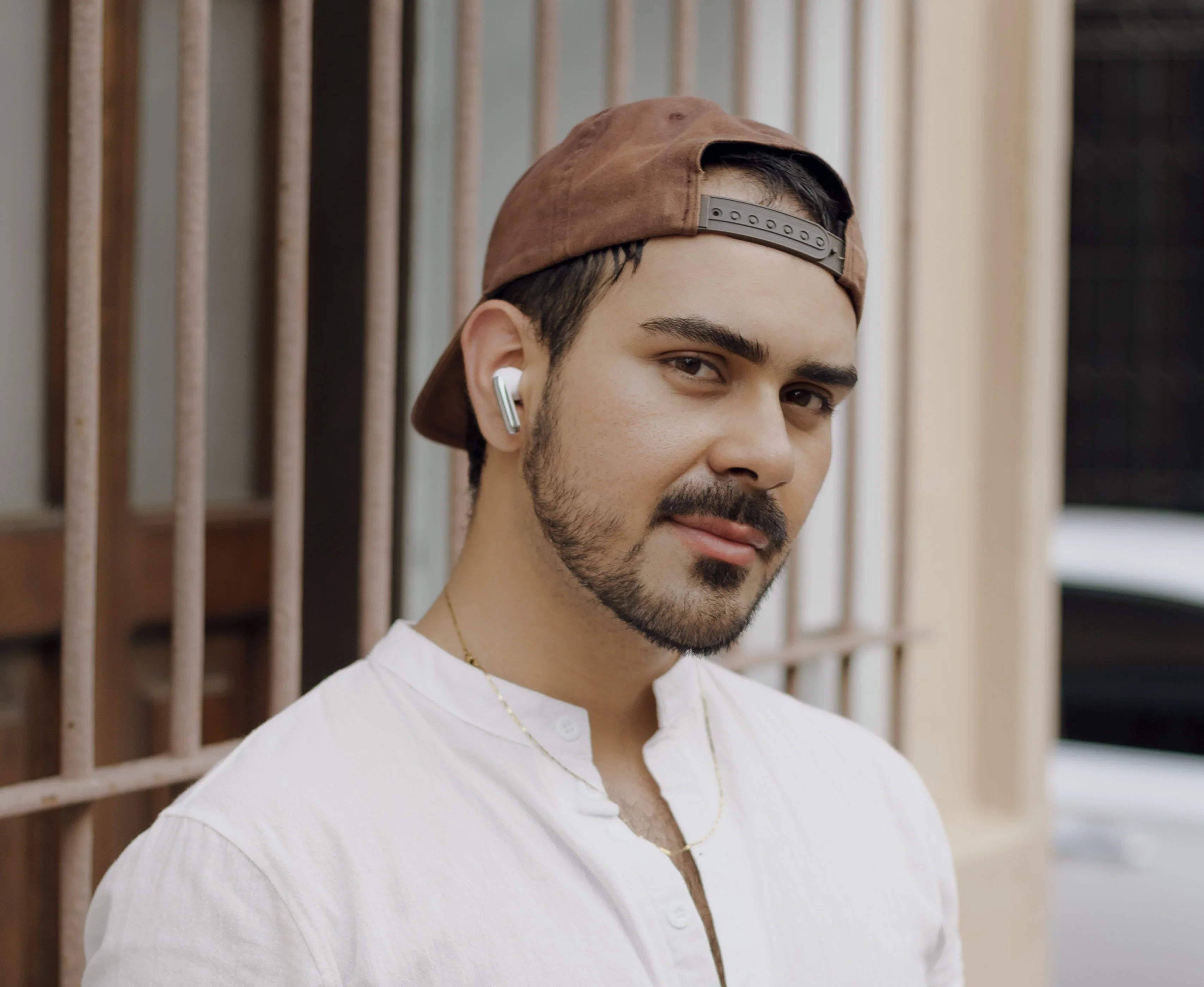 Man wearing white wireless earbuds, brown cap, and white shirt outdoors