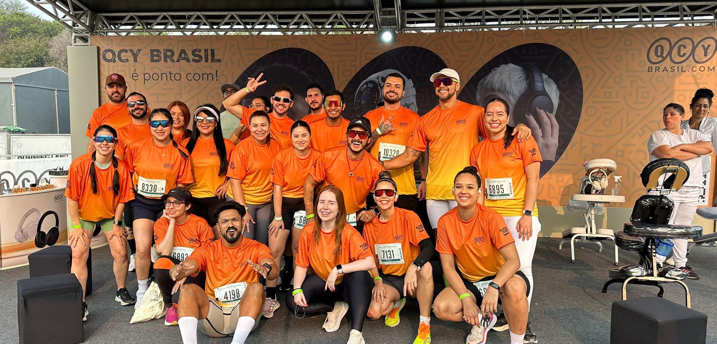 Group of runners in orange QCY Brasil shirts posing at event with wireless headphones display