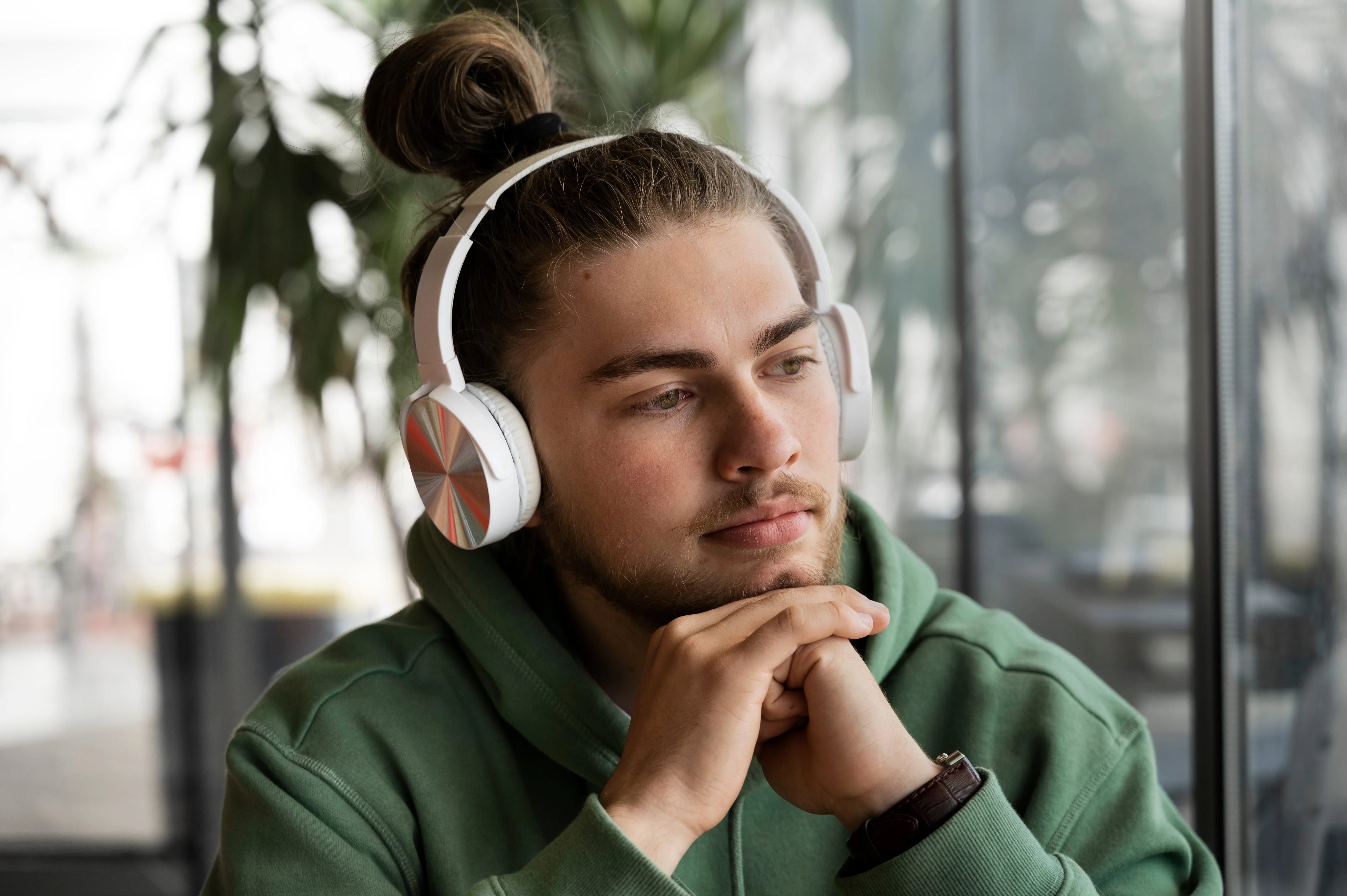 Man wearing white wireless headphones, relaxing by window with green hoodie, enjoying immersive audio experience
