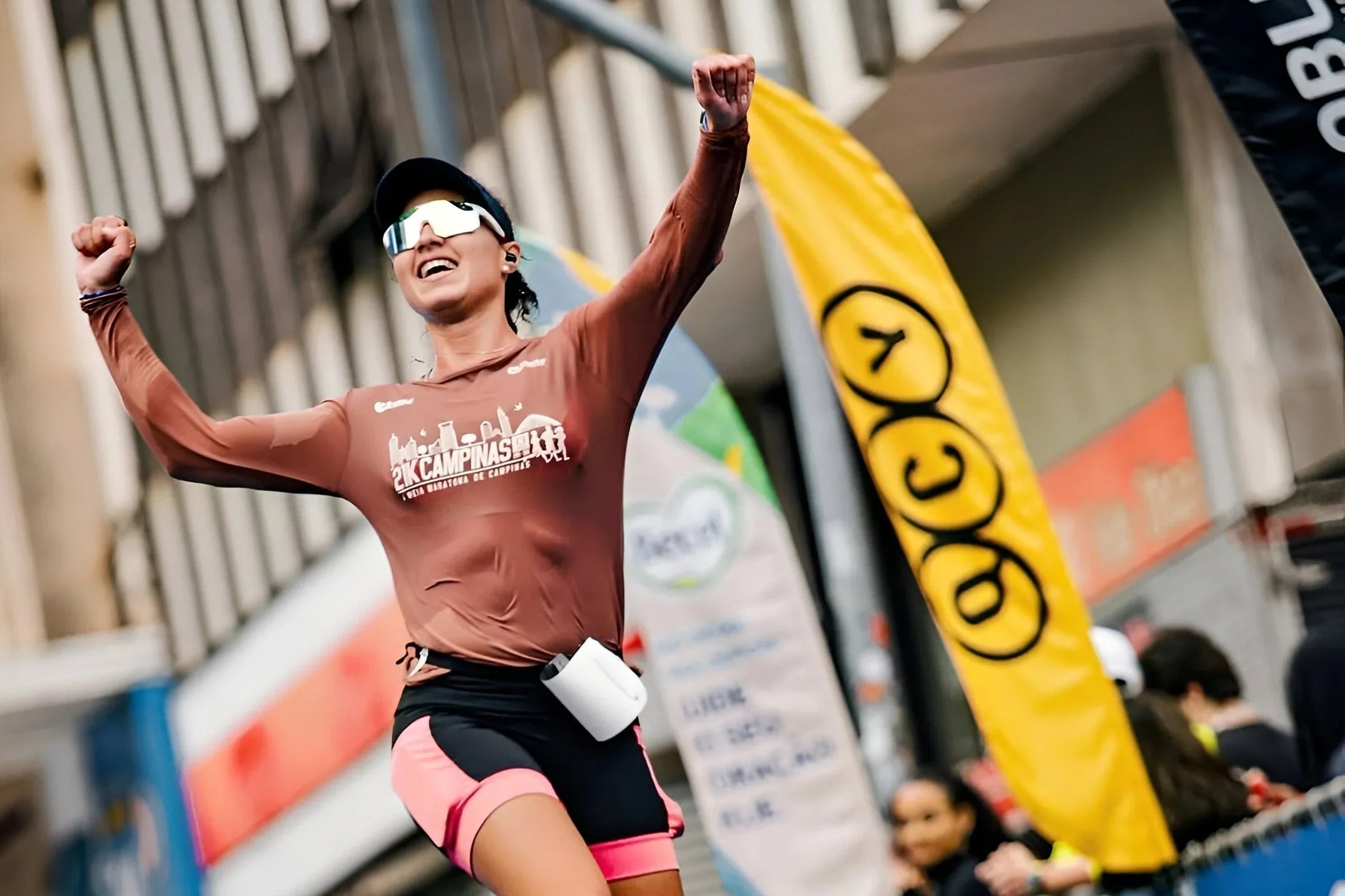 Runner celebrating finish line with wireless earbuds, wearing race gear and sunglasses at outdoor marathon event