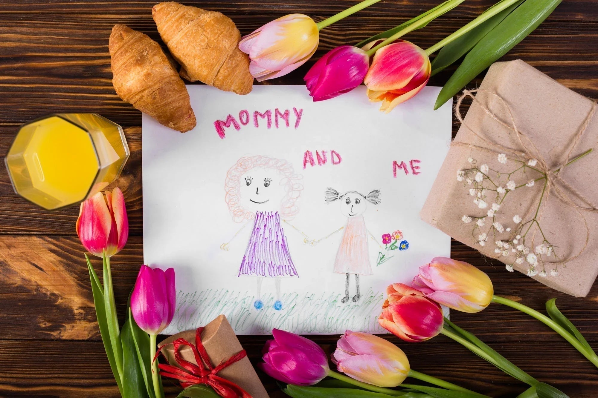 Child's drawing of mommy and me surrounded by tulips, croissants, orange juice, and gift boxes on wood table