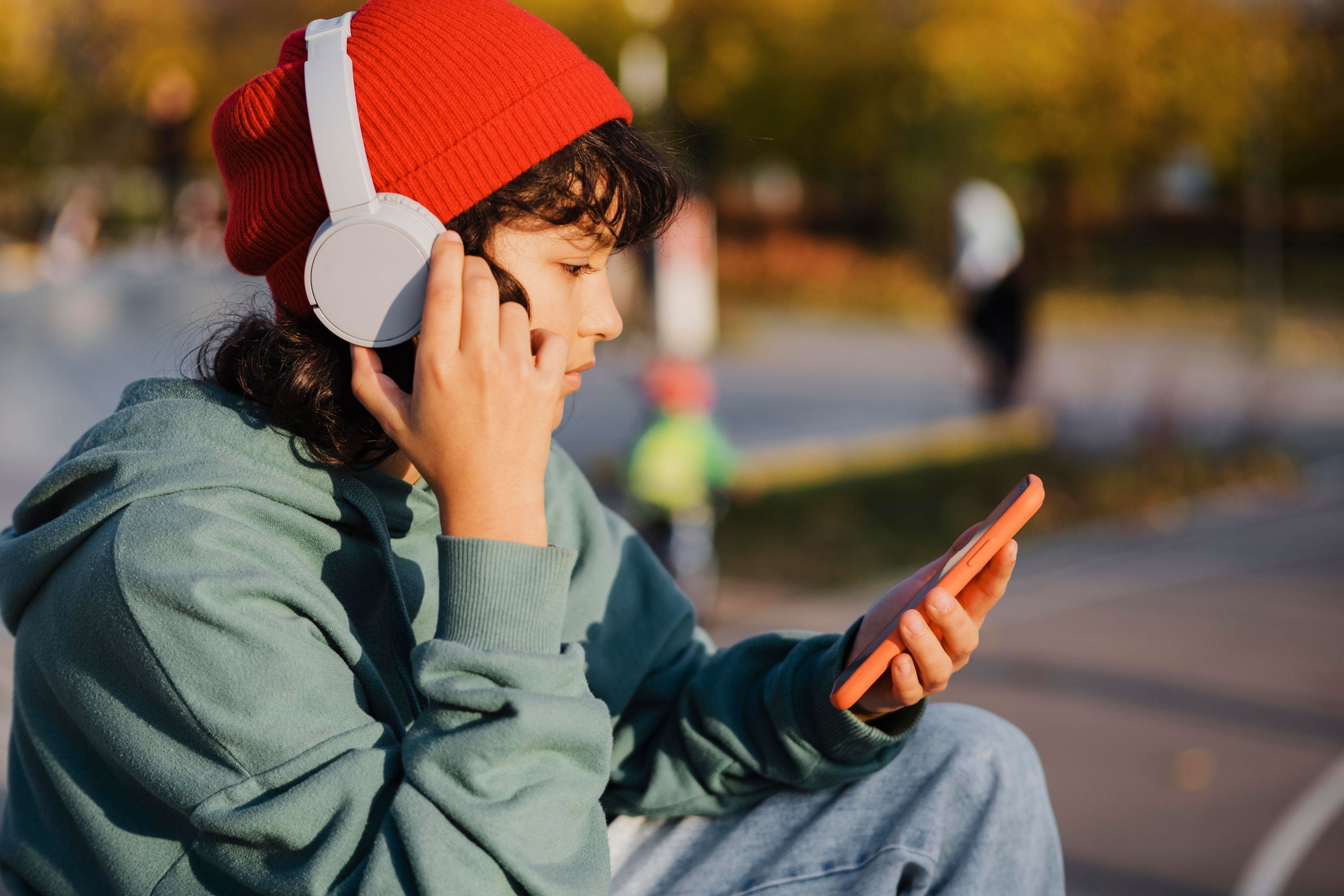 Teen wearing white wireless headphones and red beanie, using smartphone outdoors for music or calls