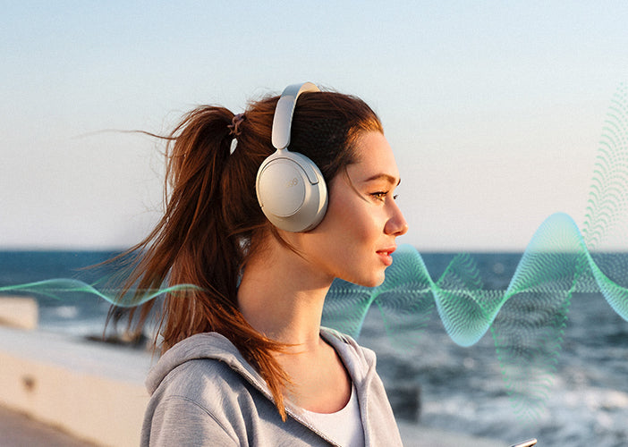 Woman wearing silver over-ear headphones by the ocean, sound waves illustrated.