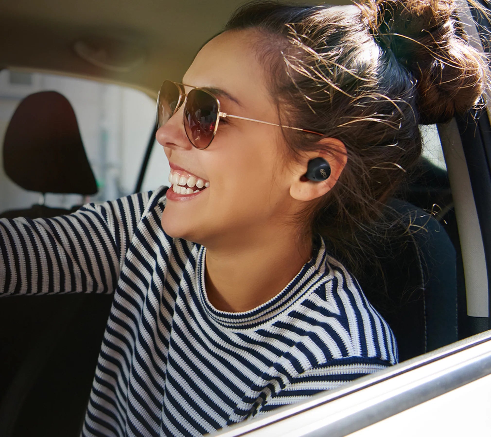 Woman in car wearing black Beats Fit Pro wireless earbuds and aviator sunglasses, smiling with sunlit hair.