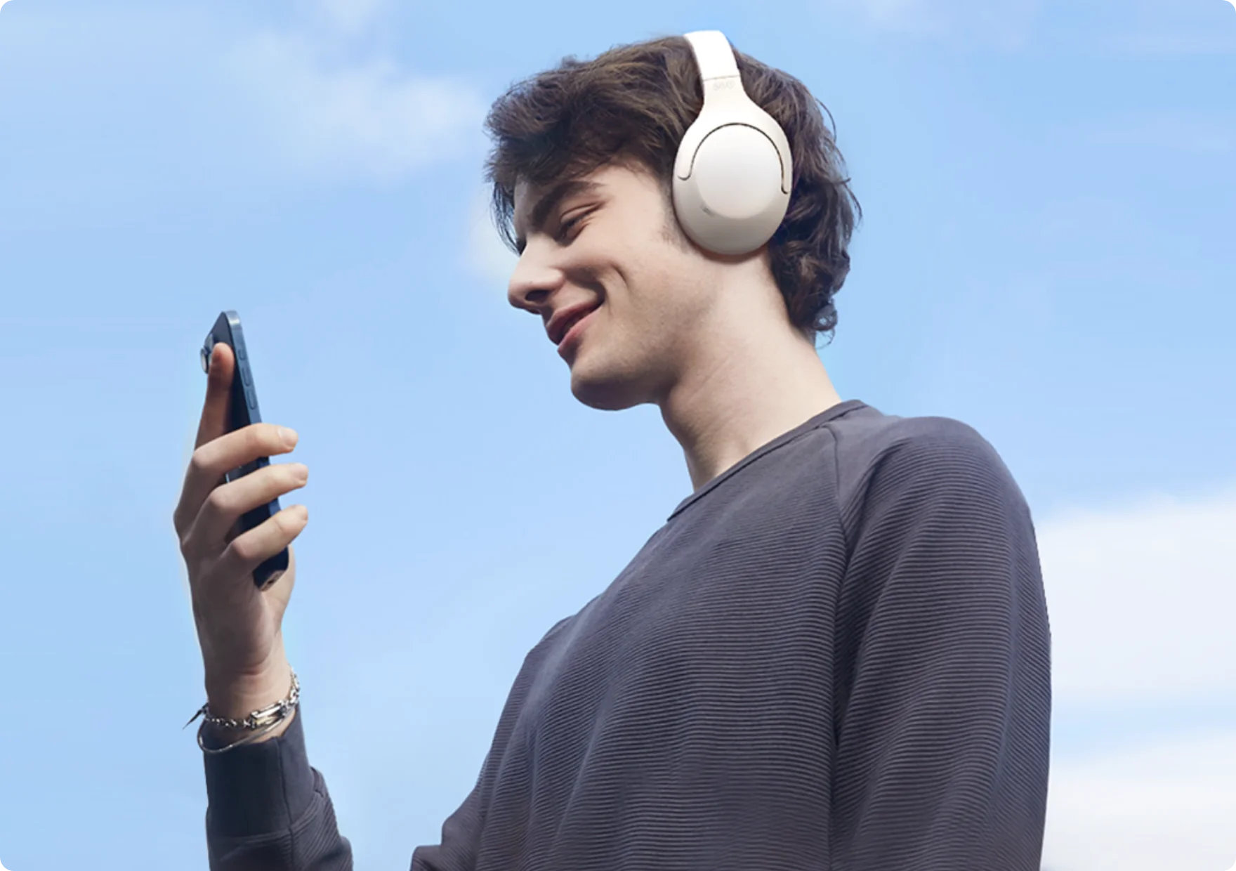 Young man enjoying music with white wireless headphones while using smartphone outdoors under blue sky.