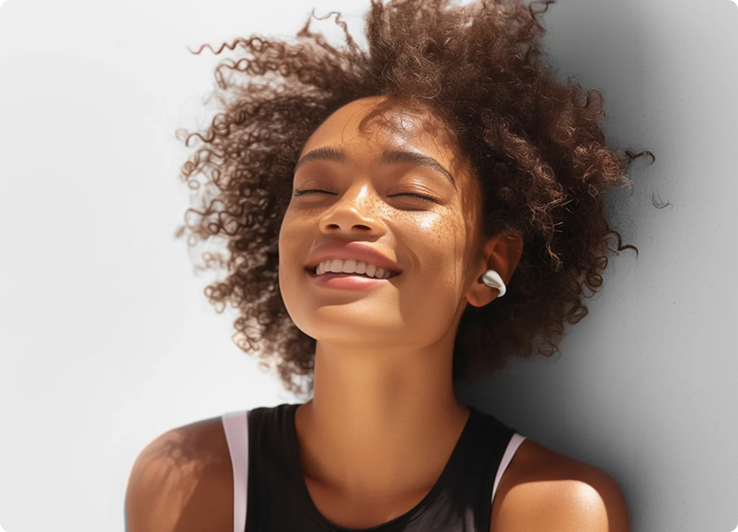 Happy woman with curly hair wearing white wireless earbuds, eyes closed, smiling in sunlight against a light wall.