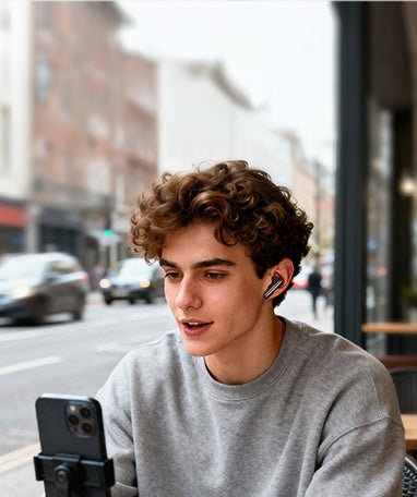 Young man with curly hair wearing wireless earbuds, listening to music on smartphone at outdoor cafe