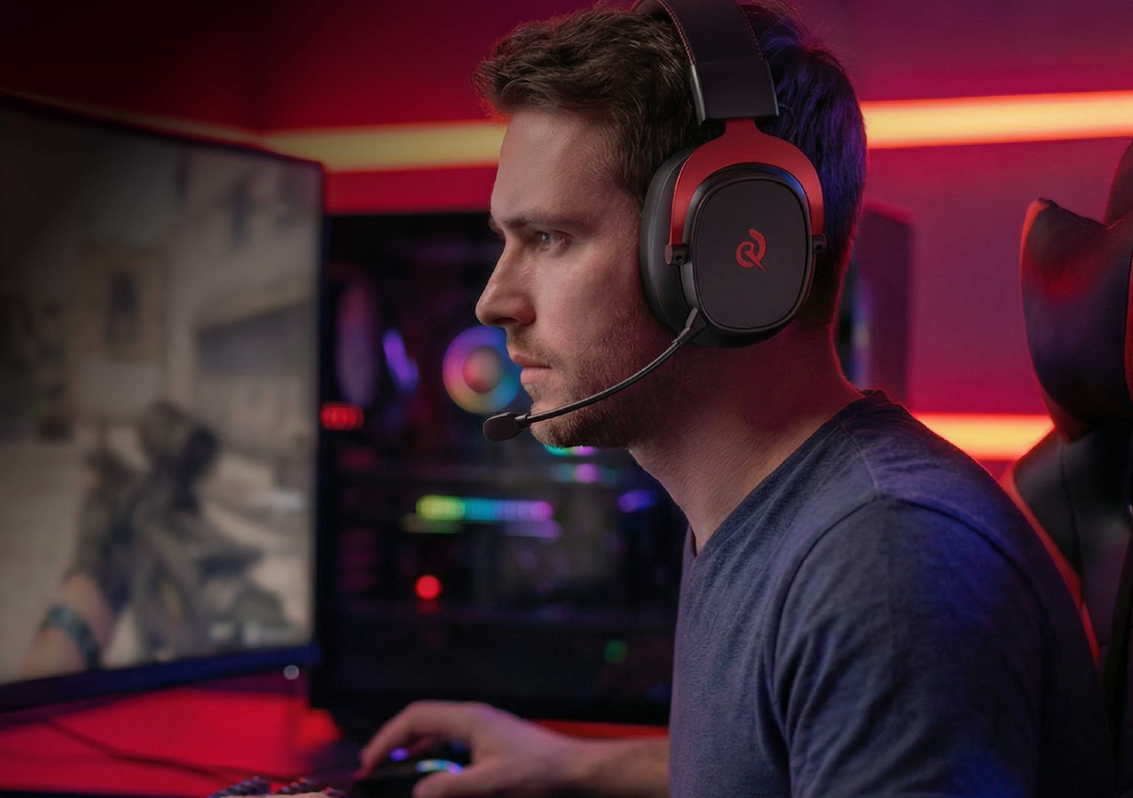 Man wearing red and black gaming headset with microphone, focused on computer screen in a vibrant, RGB-lit gaming setup.